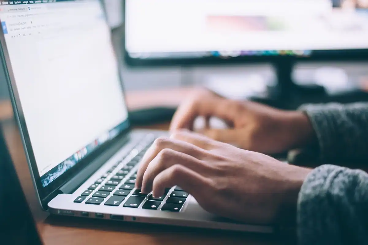 Mâinile unui dezvoltator tastează pe un laptop deschis, într-un birou luminos