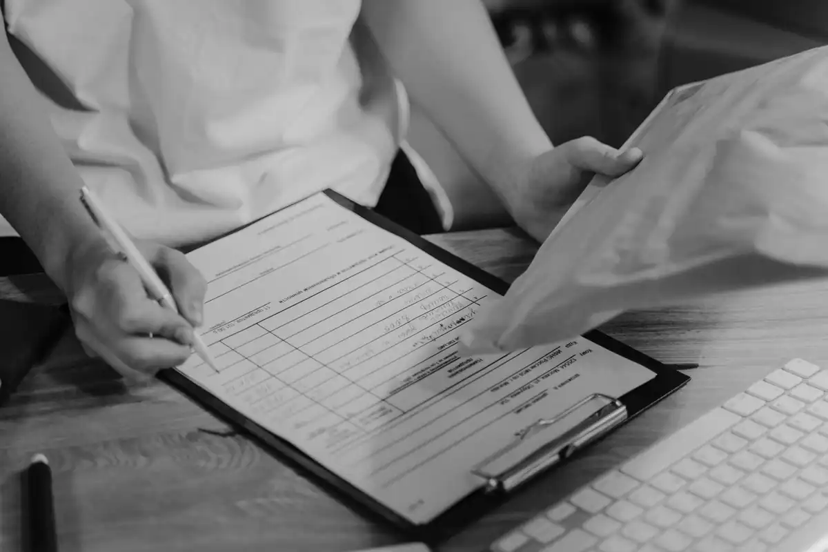Mâini completează un formular pe un clipboard, lângă o tastatură, într-o fotografie alb-negru